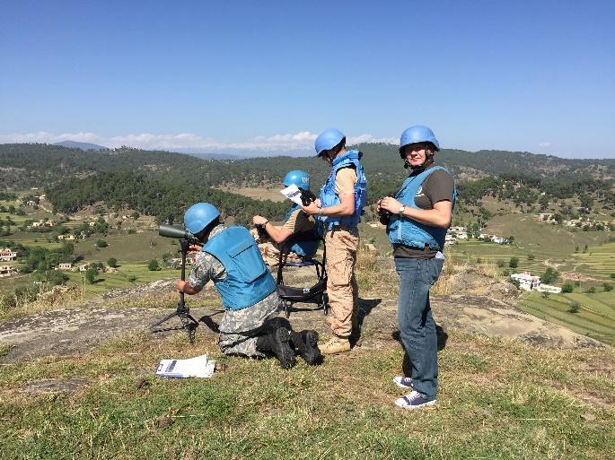 Three UN peacekeepers wearing blue helmets and vests observe terrain from a hilltop using binoculars and a tripod-mounted scope, with fields and hills in the background.
