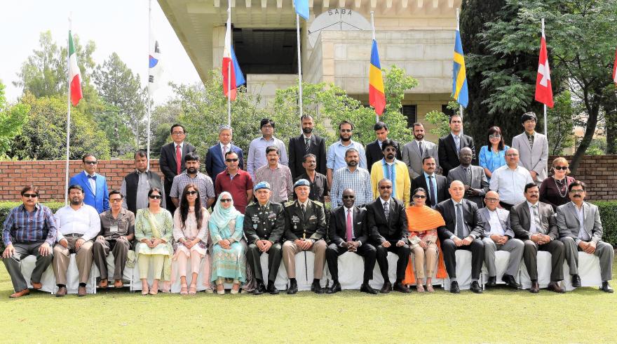 Group photo of UN civilian personnel in uniform seated and standing in rows outdoors with national flags behind them.
